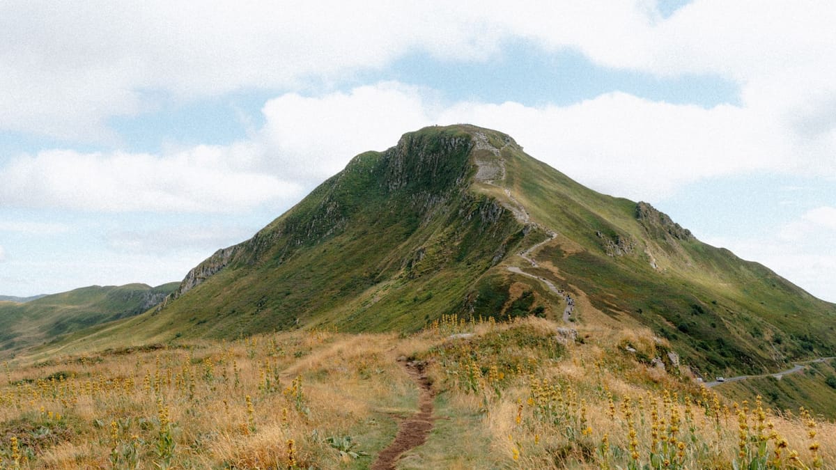 Un sentier de terre qui monte vers un sommet herbeux, sous un ciel nuageux