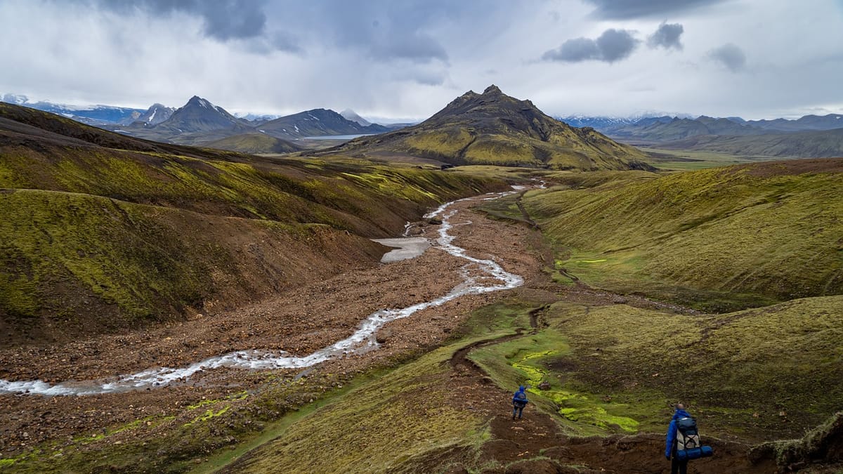 Une vallée d’herbe et de roche traversée par un torrent, avec un sentier et deux marcheurs au loin